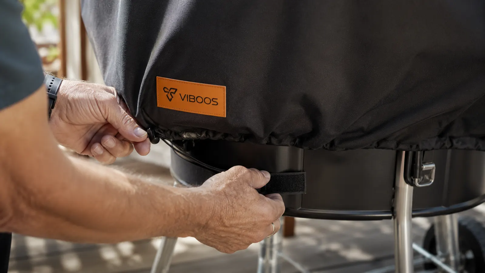 A man's hands cinching the adjustable drawstring hem of a black grill cover around the base of a gas grill