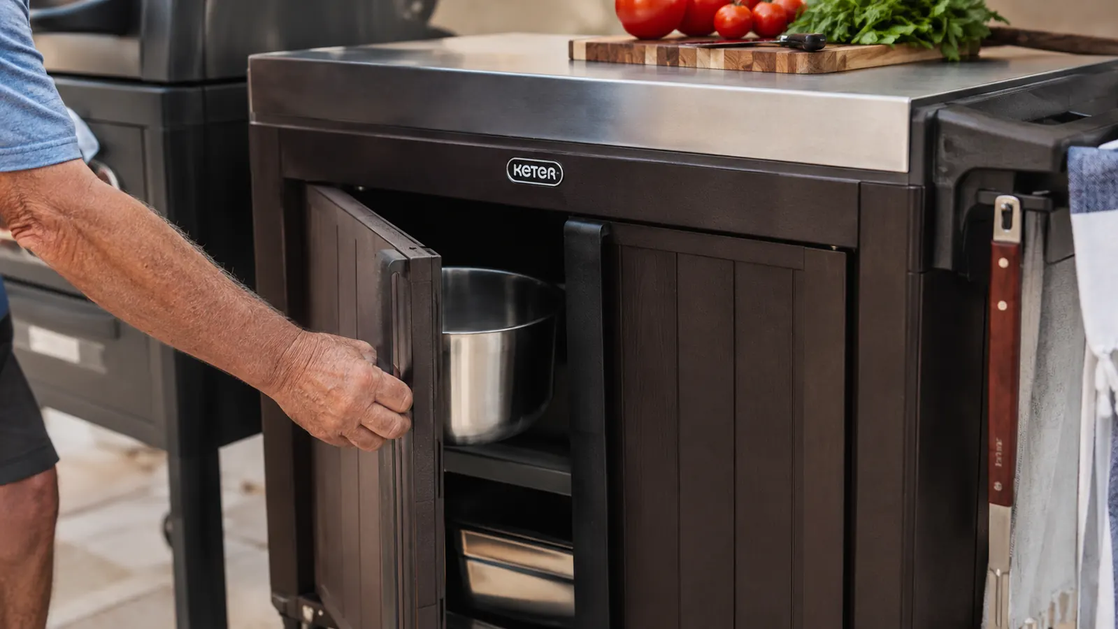 A hand opening the cabinet door of a rolling prep cart and pulling out a mixing bowl next to a lit grill