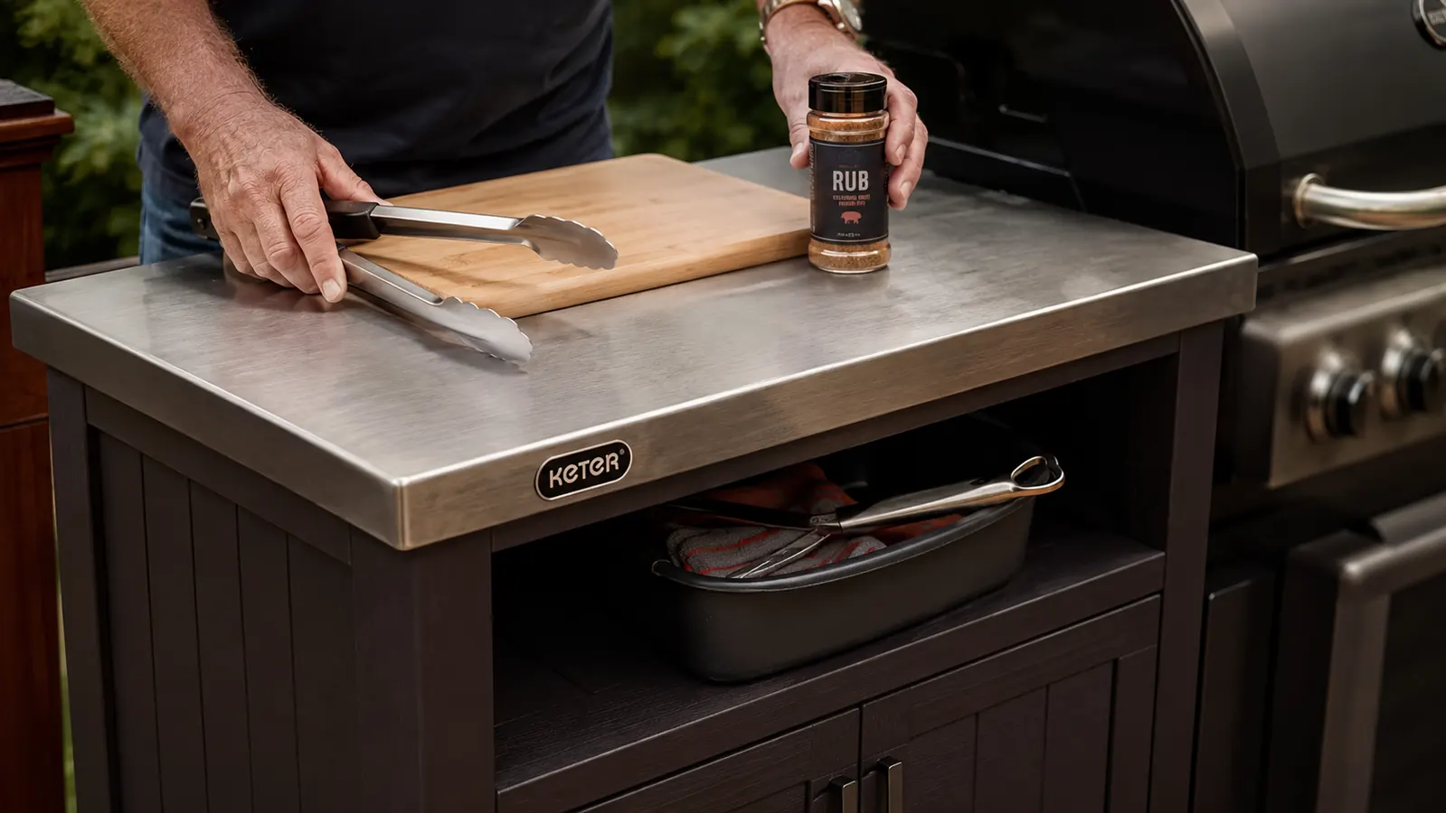 Hand setting a cutting board and tongs onto the tabletop of a dark brown Keter rolling prep cart parked beside a grill