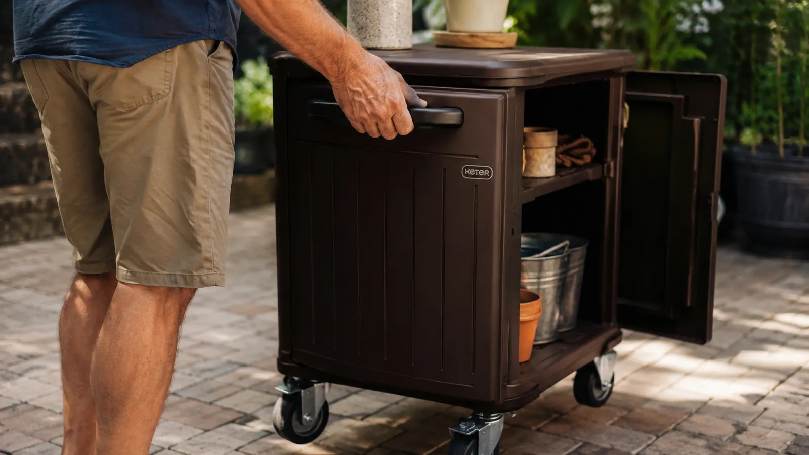 Hand rolling the Keter prep cart across a paver patio, cabinet doors and bottom shelf visible