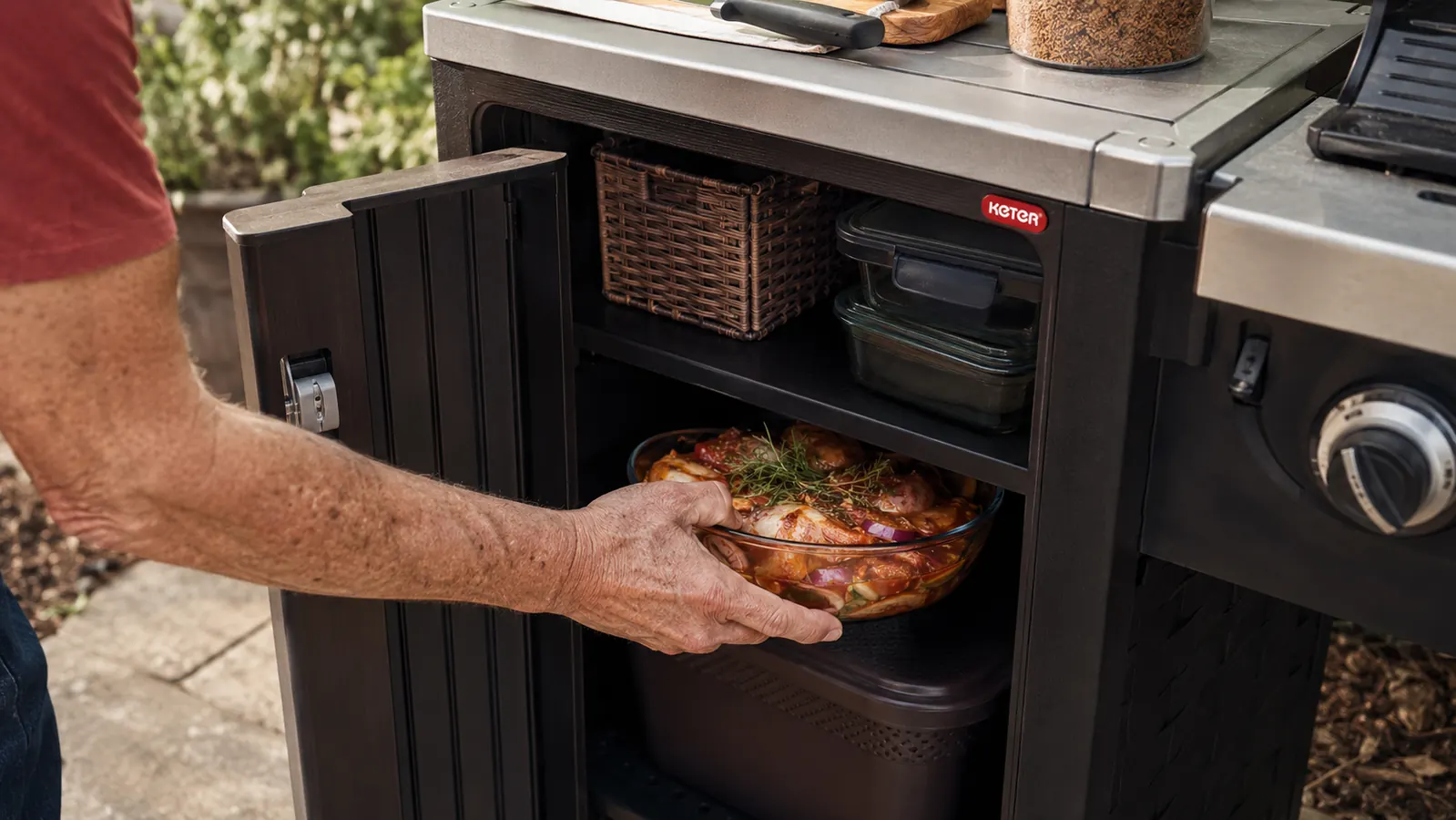 A hand pulling a stainless steel bowl of marinated chicken out of the enclosed cabinet of a rolling prep cart next to a grill