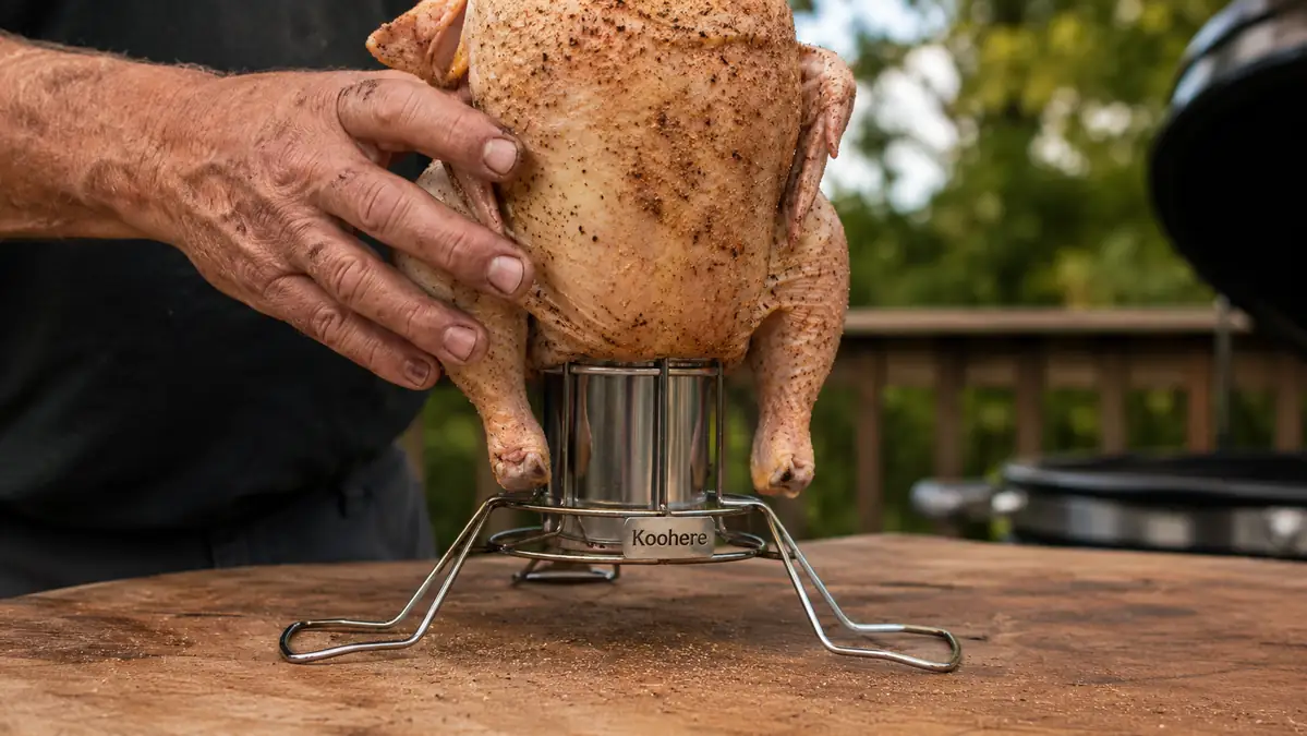 A hand lowering a seasoned whole chicken down onto a stainless steel vertical holder with its liquid reservoir already filled