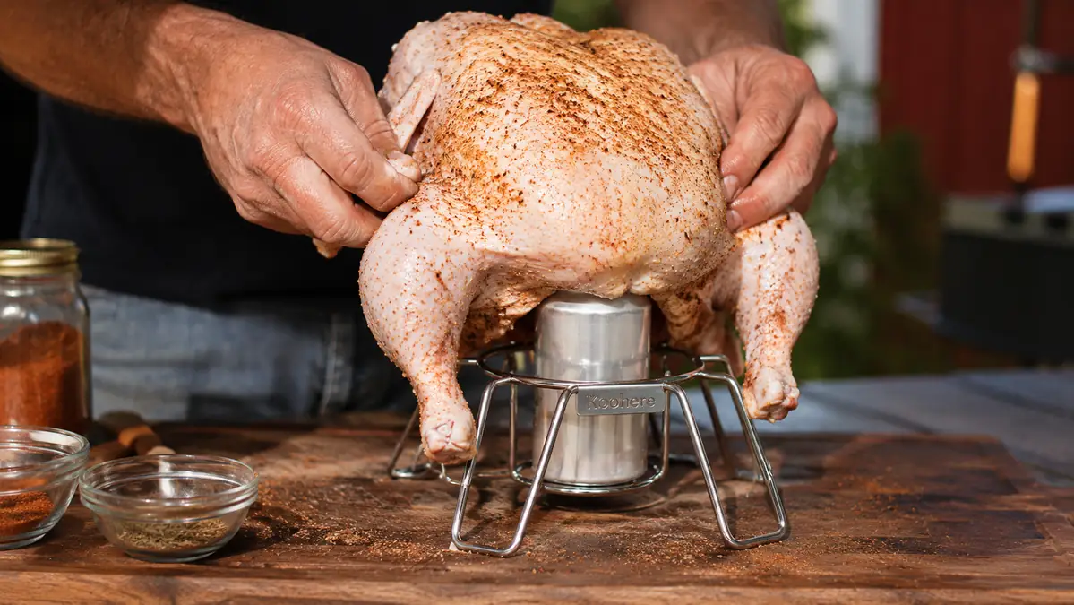 Hand setting a whole raw chicken down onto a stainless steel Koohere vertical chicken stand with beer can holder on a kitchen counter