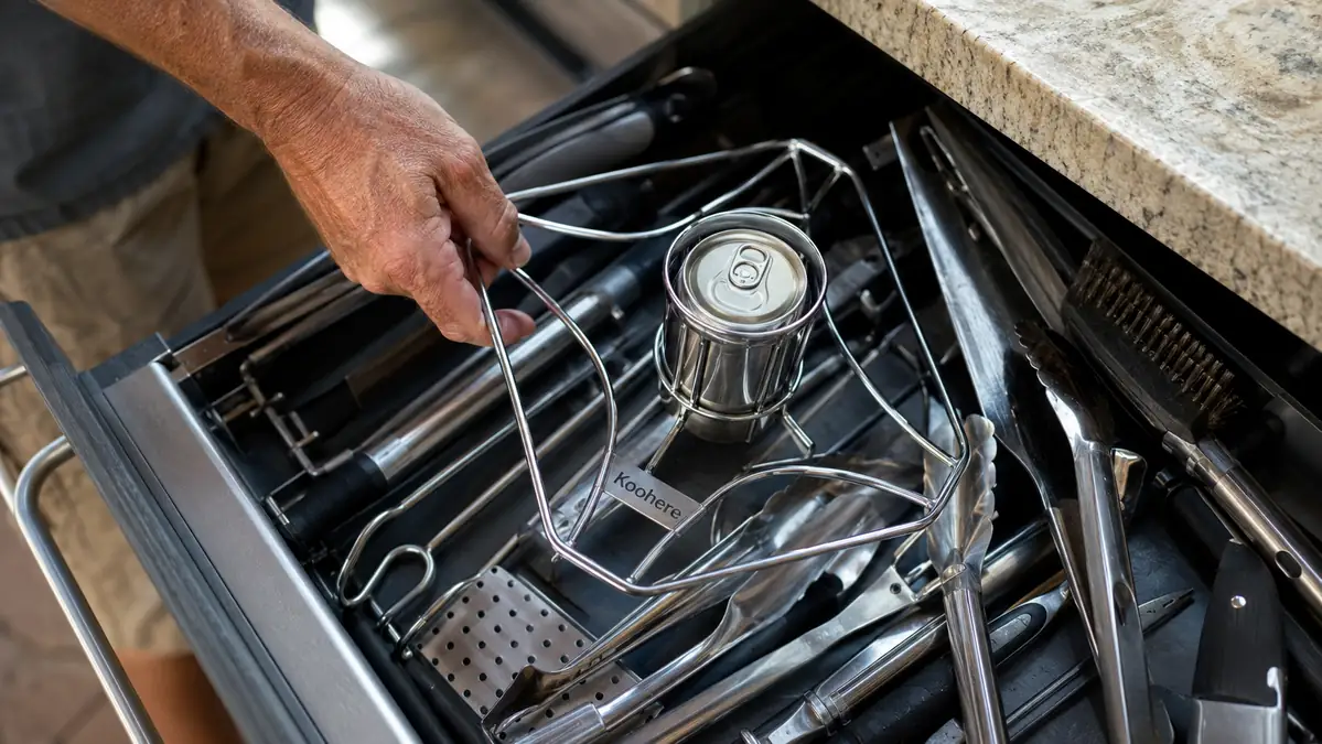 A cluttered kitchen utensil drawer with a stainless steel chicken holder wedged in among tongs and spatulas