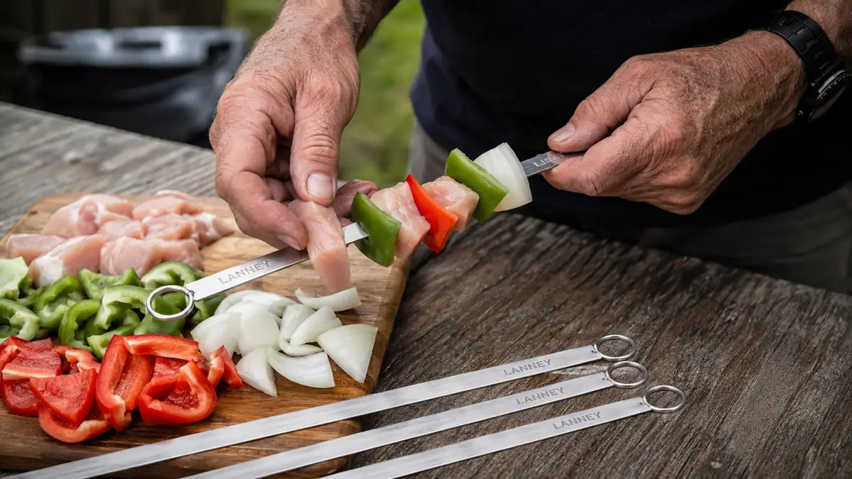 Hand threading chicken and pepper chunks onto a flat stainless steel LANNEY skewer over a cutting board