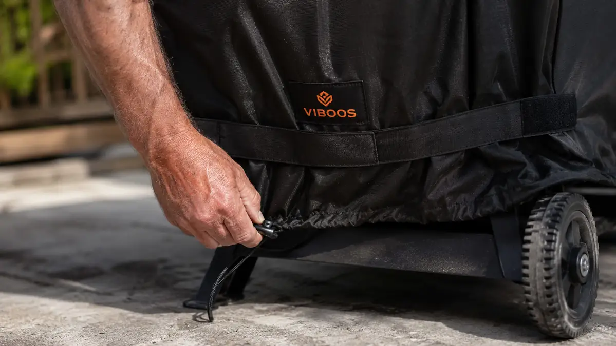 A hand pulling the drawstring hem of a black VIBOOS grill cover snug around the base of a gas grill before a storm