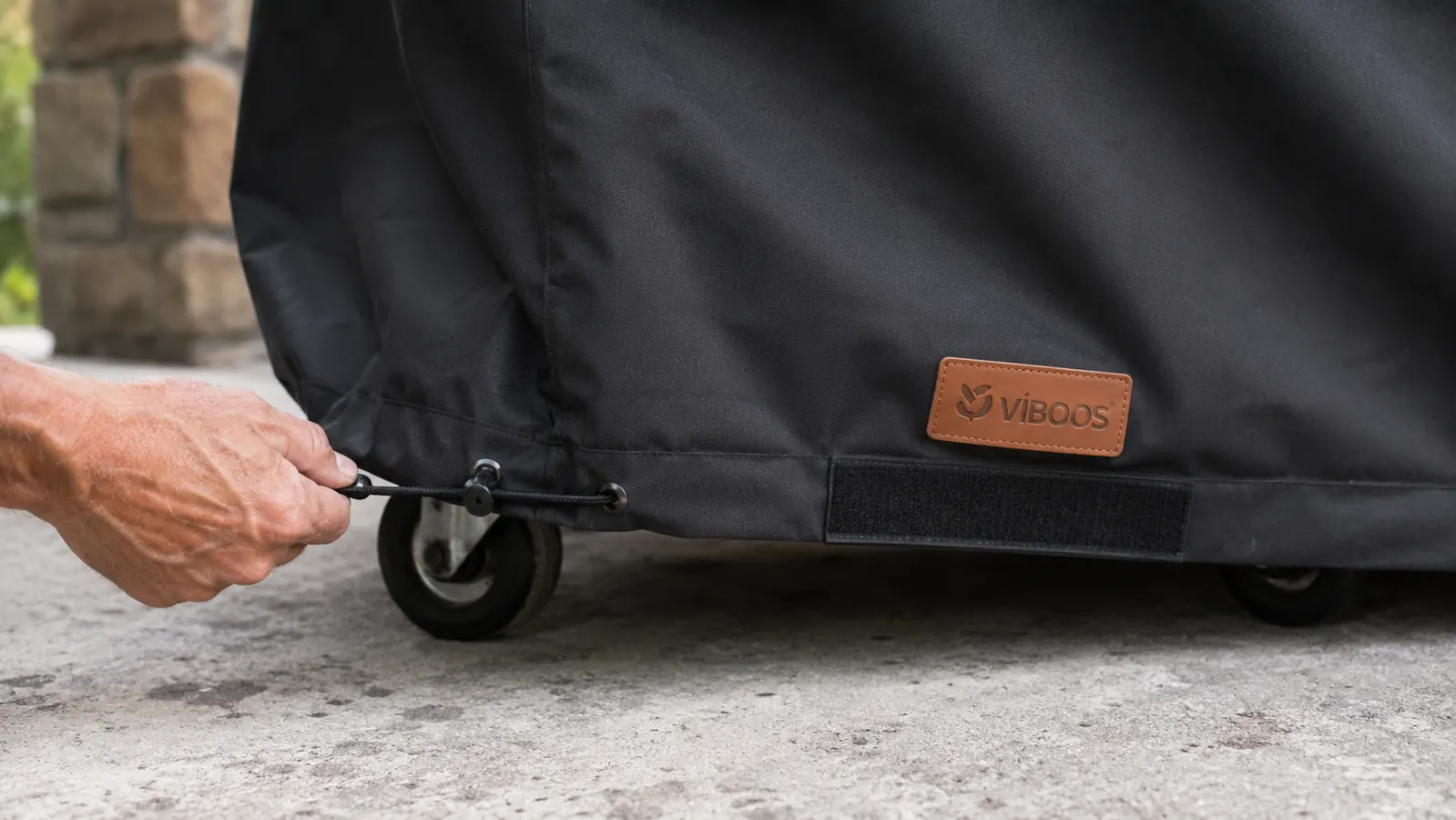 Hand cinching the adjustable hem drawstring on the bottom of a black VIBOOS grill cover around the base of a gas grill