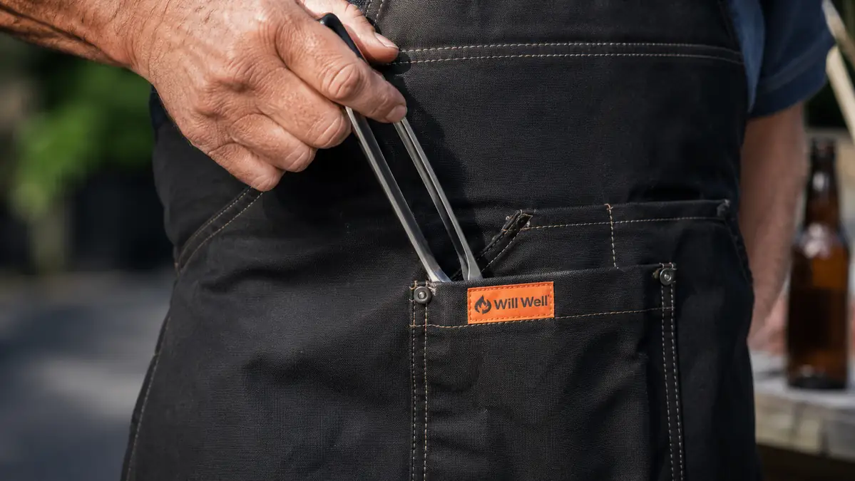 A hand reaching into the front pocket of a black Will Well apron to pull out a pair of stainless tongs