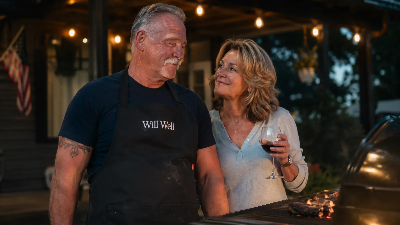 A husband and wife standing together near a grill at dusk, the man wearing a black apron with visible grease marks near the pocket
