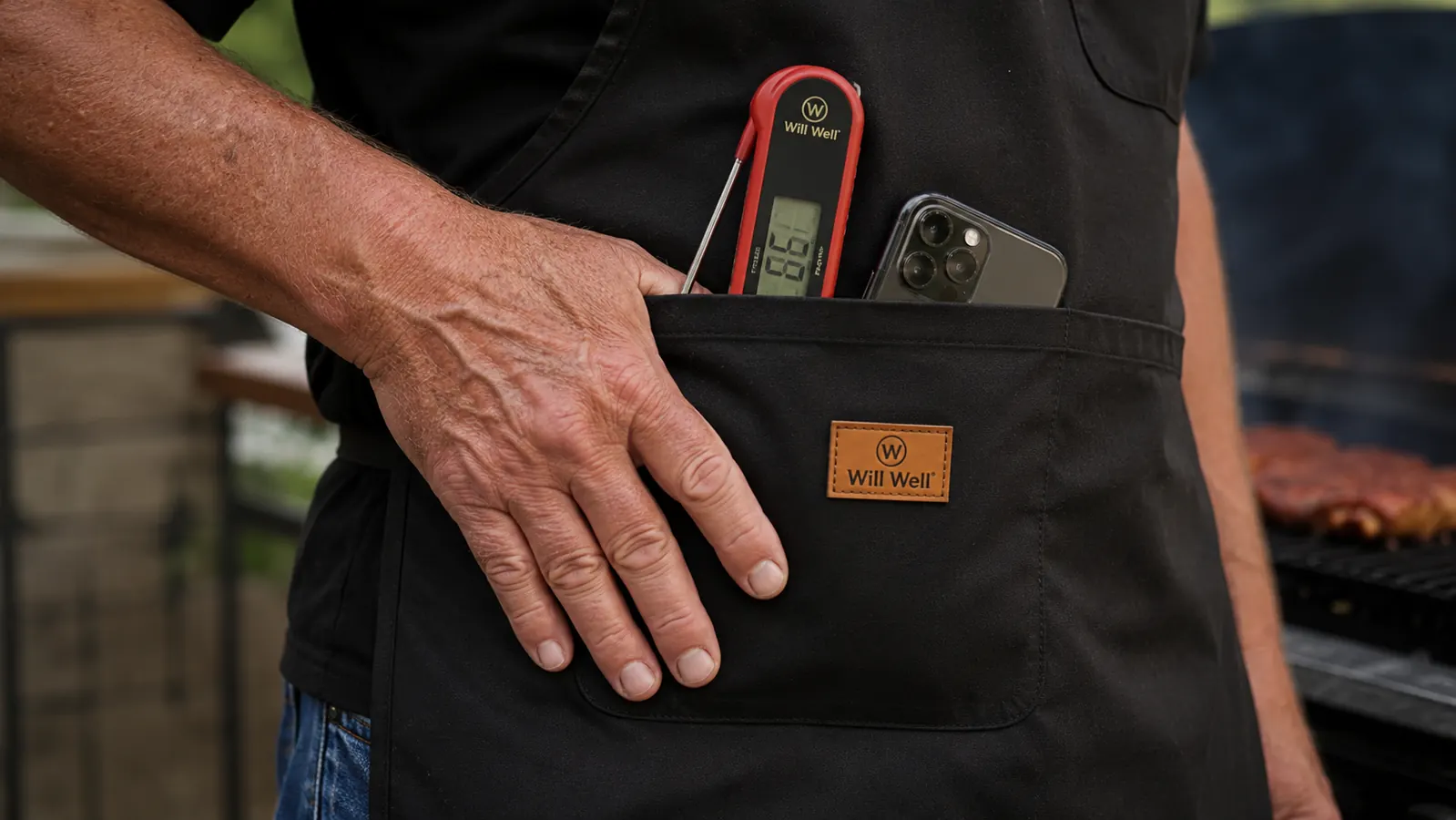 A hand pressing down on a bulging front pocket of a black apron loaded with a thermometer and a phone