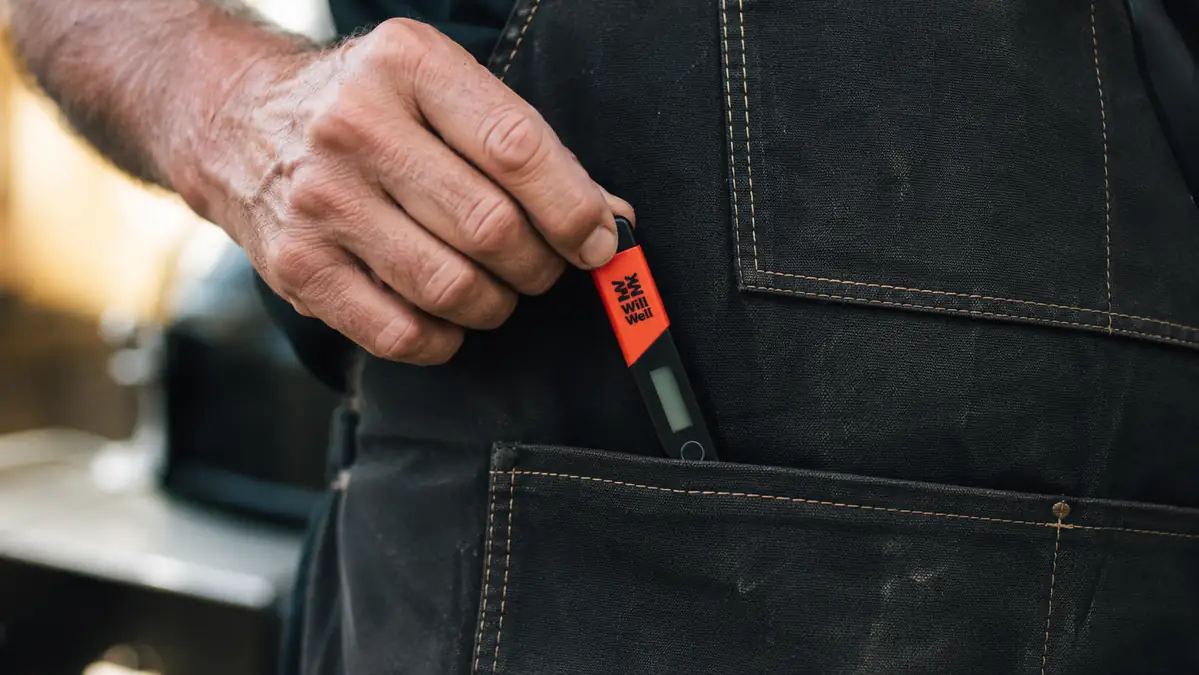 A hand reaching into the front pocket of a black Will Well apron to pull out an instant-read thermometer
