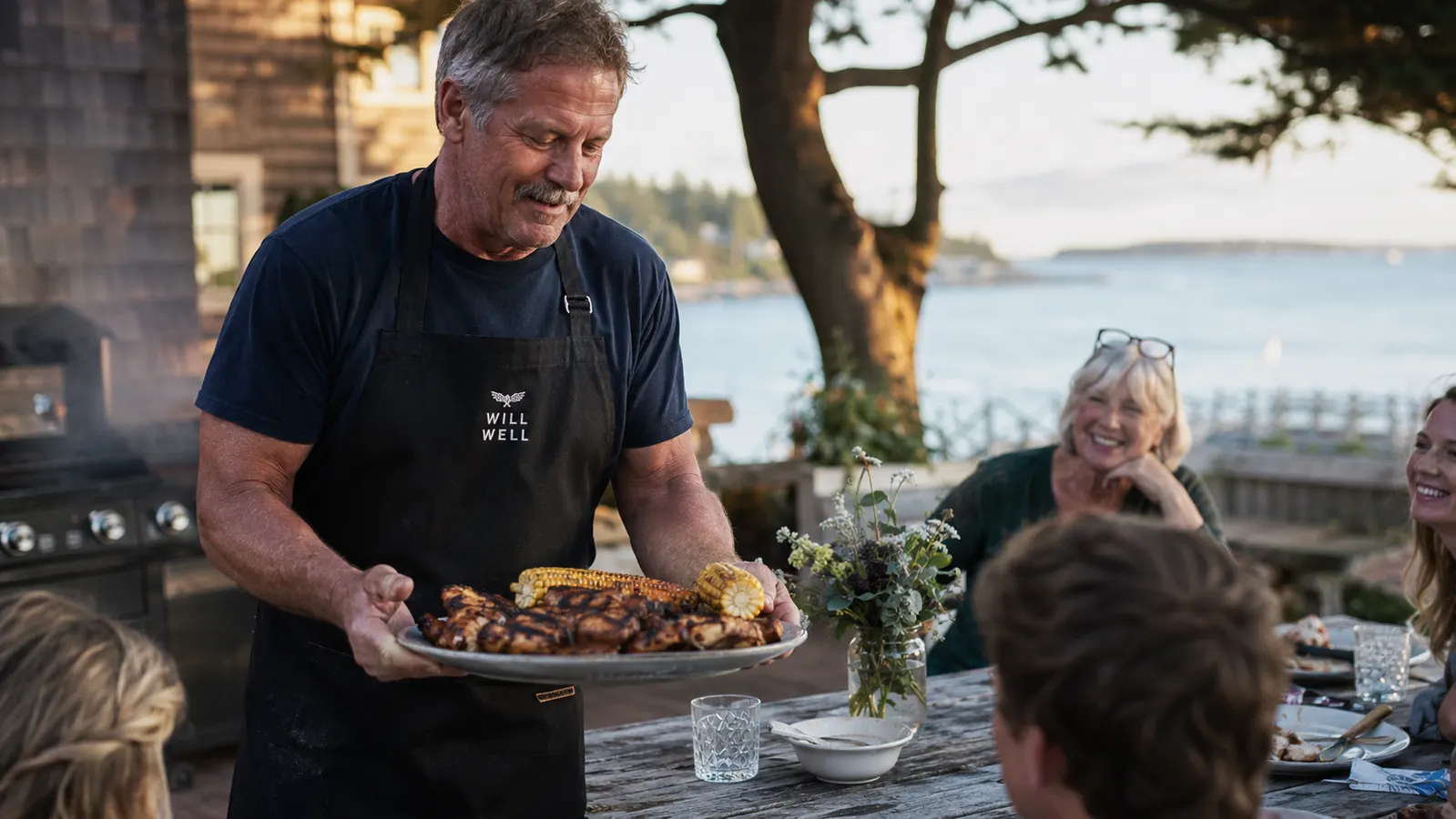 A family gathered around a picnic table while a man in a black apron carries a platter of grilled food to the group